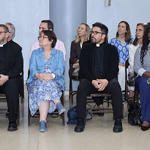 Clergy and attendees sit in chairs, listening attentively.