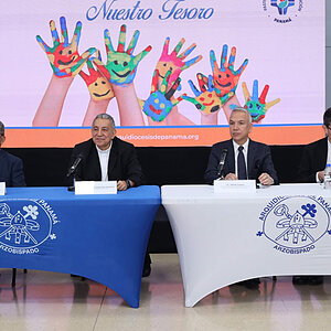 Four men in suits and clerical attire sit at tables, with a screen displaying colorful painted hands behind them.