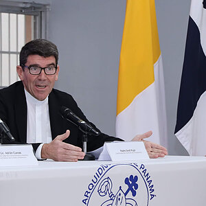 Man in black suit and white clerical collar speaks, gesturing with his hands.