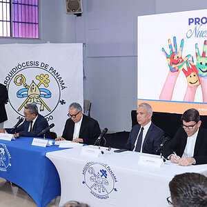 Five men in suits sit at tables, with one speaking at a podium before a screen displaying colorful hands.